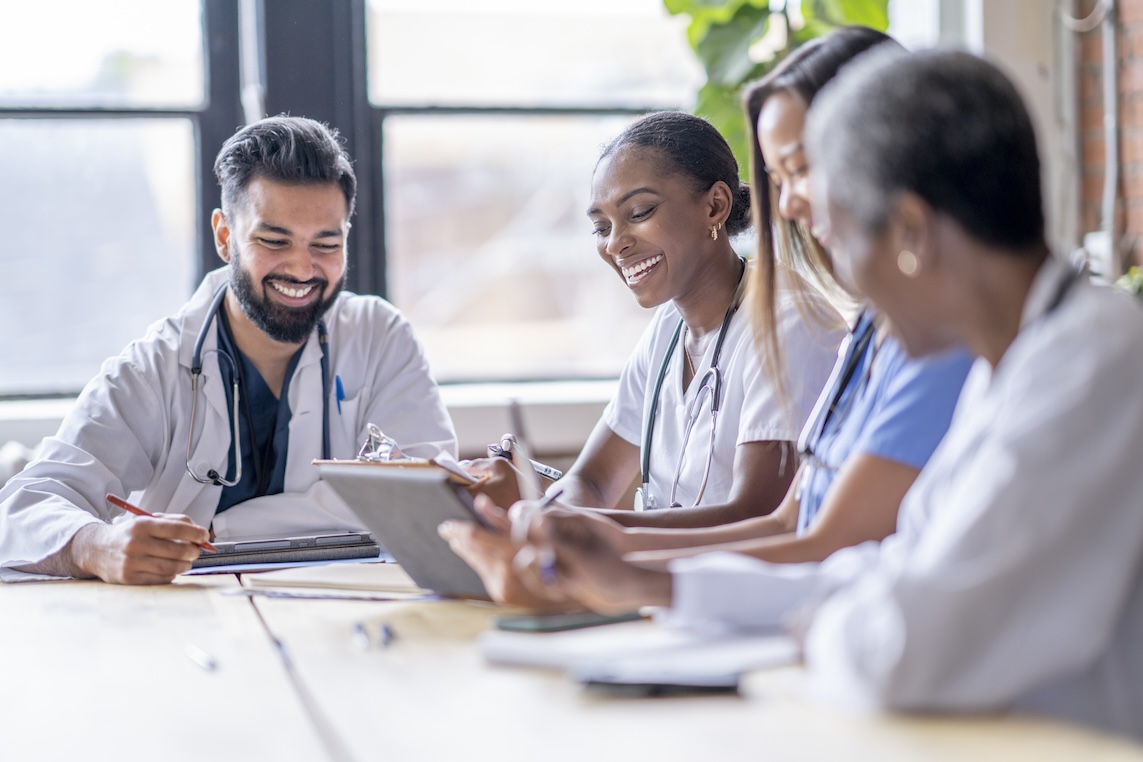 A small group of four medical professionals sit around a boardroom table as they meet to discuss patient cases.  They are each dressed professionally in scrubs and lab coats as they focus on working together.